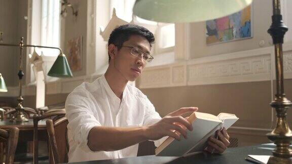 跟踪拍摄年轻的亚洲男学生在大学阳光图书馆沉思读书的镜头