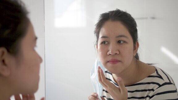 年轻的亚洲成年女性担心戴口罩后出现的皮肤痤疮或丘疹在镜子中显示出下巴和脸颊的肿胀、斑点、疤痕和皮肤过敏