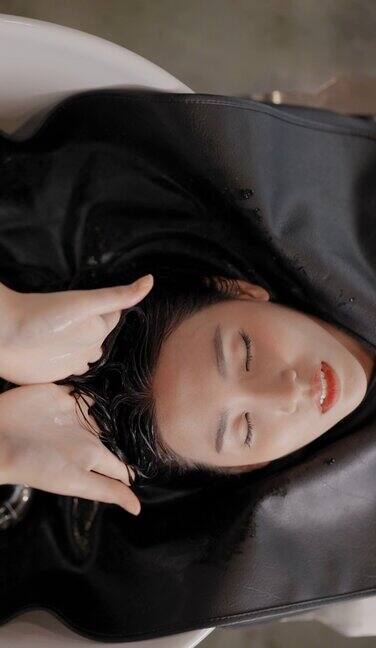 俯视图迷人的年轻亚洲女孩躺在一个沭浴盆微笑和放松闭上眼睛而理发师温柔地按摩客户的头和洗涤