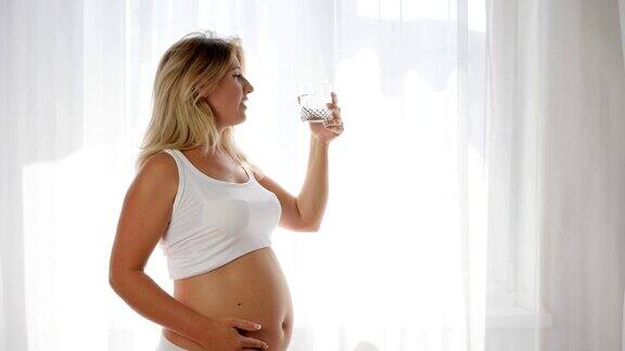 肖像孕妇饮用矿泉水在玻璃和摩擦裸露的腹部靠近窗户