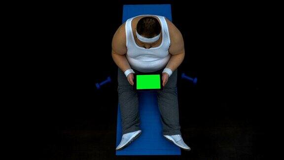 肥胖的男性坐在健身房的平板电脑上滚动阅读健康生活方式的文章