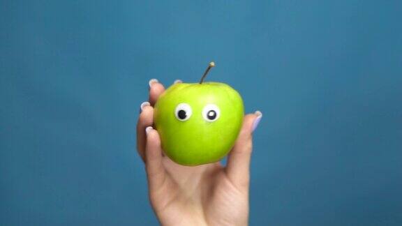绿苹果与眼睛在一个女人的手特写苹果在蓝色背景上摇动眼睛缓慢的运动