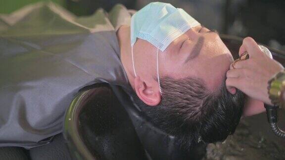 戴着保护面罩的亚洲华人男性在美发沙龙里闭着眼睛躺着洗头