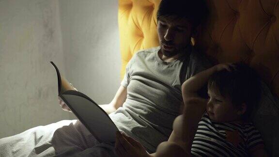 晚上睡觉前年轻的父亲和小儿子躺在家里的床上看童话书
