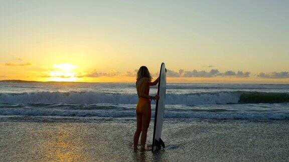 美丽的年轻女子在泳衣站在海里同时持有冲浪板大海浪和日落的背景