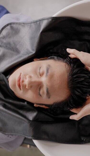 俯视图英俊的年轻亚洲男子躺在一个沭浴盆微笑和放松闭上眼睛而理发师温柔地按摩客户的头和洗涤