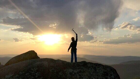 一名年轻快乐的雄性登山者站在山顶成功举起手臂手持冰斧享受日落时分的美景