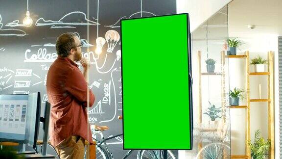男性开发者看着带有绿色屏幕模型的交互式白板他捋着胡子思考解决问题的方法在背景创意办公室与黑板墙与项目计划绘制
