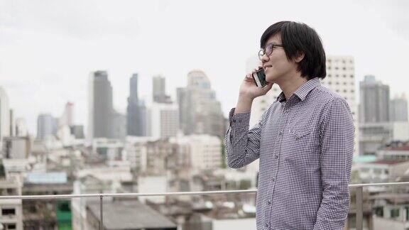 年轻的亚洲商人站在写字楼屋顶露台上用智能手机谈生意背景是城市景色商务沟通和企业理念