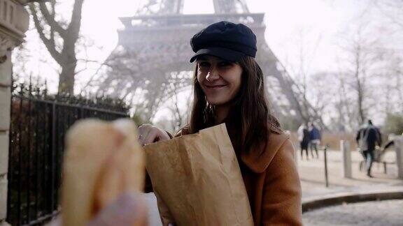 美丽而快乐的欧洲女人走在埃菲尔铁塔附近慢动作地把一块法式长棍面包递给摄像师