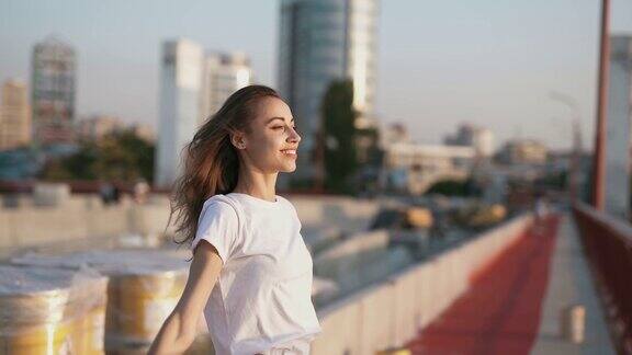 身穿白色t恤的年轻漂亮女子高高举起双手站在城市背景上的桥上快乐地微笑着欣赏着宁静的日落向前看平静和放松
