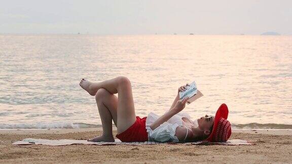 美丽的女人躺在沙滩上幸福地在海滩上放松地看书日落