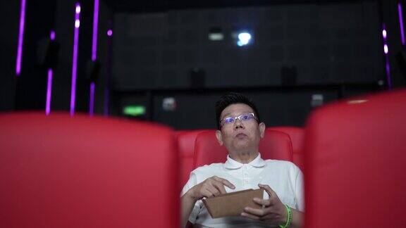一名亚裔中国男子坐在空荡荡的电影院的红色座位上一边吃一边看电影