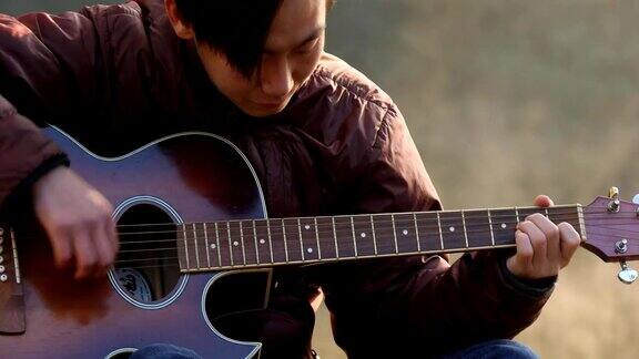 英俊的年轻亚洲男子在森林河海岸弹吉他在日出黎明薄雾的背景