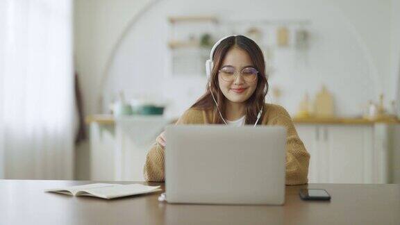 年轻的亚洲妇女戴着眼镜和耳机在家里的笔记本电脑上工作在家办公视频会议视频通话学生在线学习