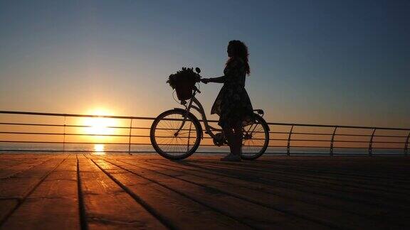 年轻迷人的女人的剪影穿着复古复古的自行车和鲜花走在海边的木堤岸在日出或日落浪漫的旅行概念4k