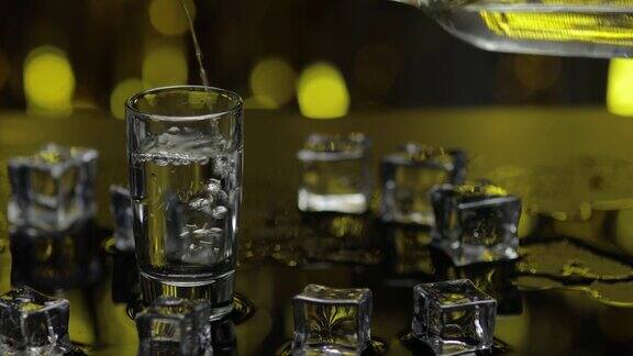 酒保将冰冻的伏特加从瓶子倒入小酒杯冰块映衬着闪亮的金色派对背景