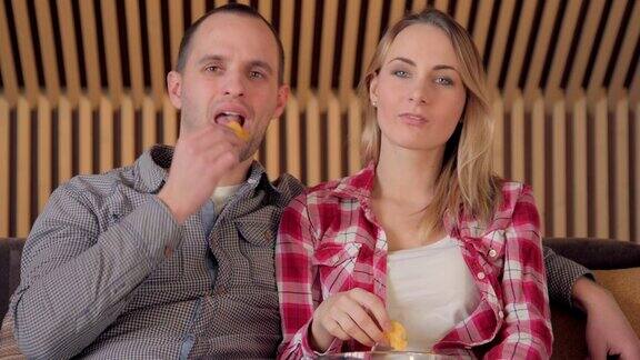 年轻夫妇坐在客厅的沙发上看电视一边吃薯条
