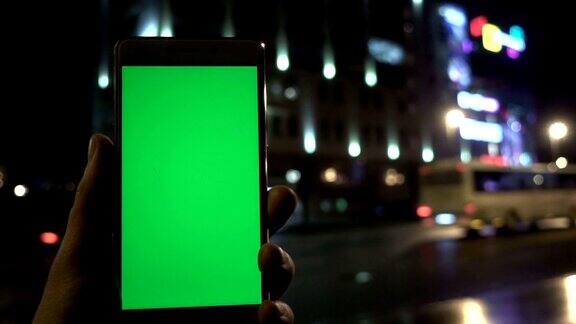 男子手握白色智能手机在夜城灯光散景的背景下绿色的屏幕