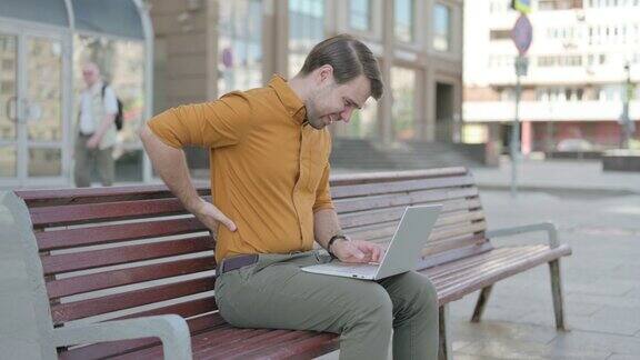 一名背部疼痛的年轻人坐在户外长凳上使用笔记本电脑