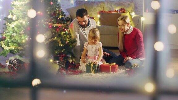 透过白雪的窗户看快乐的爸爸妈妈和女儿坐在圣诞树下女儿打开礼物