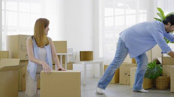快乐的年轻夫妇打包纸箱准备搬到新家或公寓漂亮的年轻女子包装纸箱和微笑而她的丈夫持有和重新安排纸箱到客厅的角落
