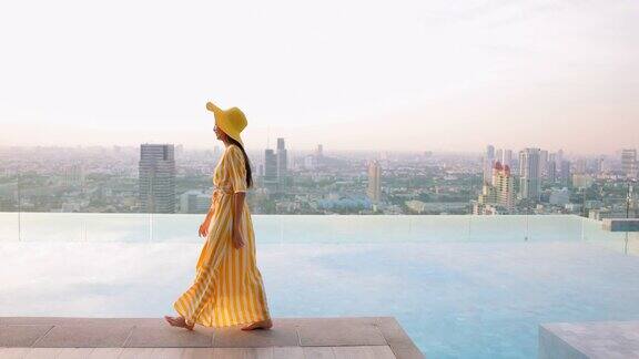 美丽的女人走在池边穿着黄色的长裙和黄色的帽子女人们漫步在无边的游泳池旁伴着夕阳的城市风光