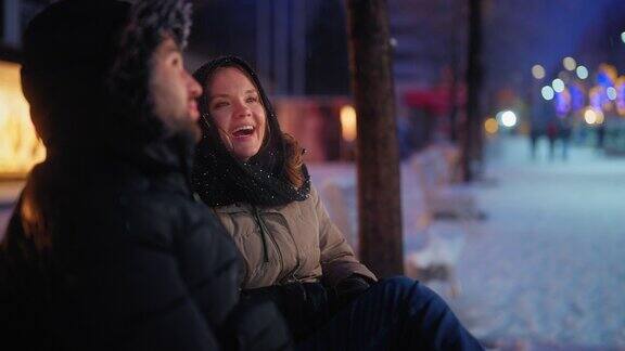 年轻的情侣约会和坐在长椅上聊天而在城市的雪晚上