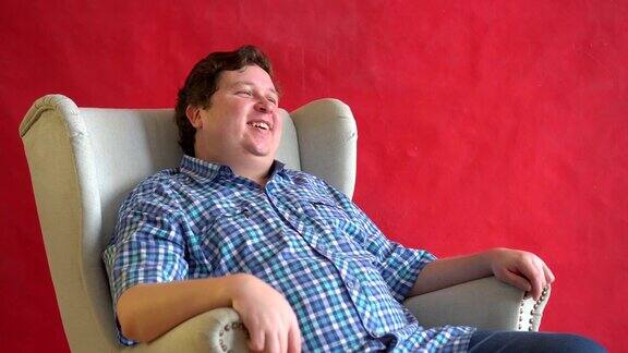 年轻的胖子格子衬衫坐在椅子上大声笑孤立在红色的背景-有趣