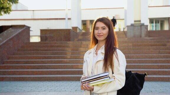 开朗的女学生抱着书背着背包站在学校大楼外对着镜头微笑现代教育大学学费和助学金学生和留学概念