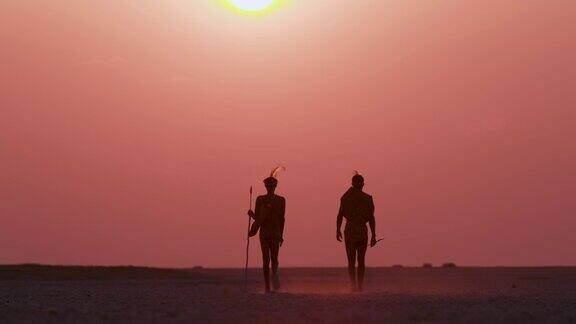 在博茨瓦纳的Makgadikgadipan上两名圣布须曼人走向夕阳下的红色天空的后视图