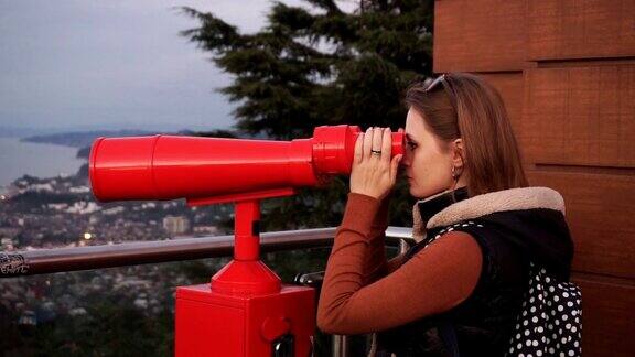 旅游:年轻的女游客在日落时用投币望远镜看城市中景手持慢镜头60帧秒