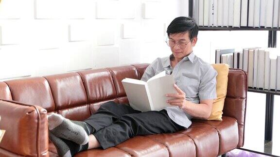 年长的亚洲男人在家里客厅的沙发上看书亚洲老人正在放松和幸福读杂志