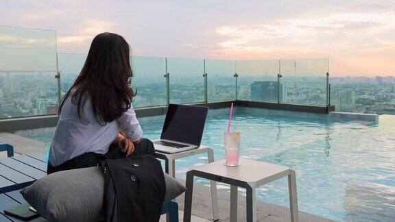 亚洲妇女在工作日缺乏精力疲惫的女商人感到无聊坐在摩天大楼的泳池边拿着笔记本电脑和荔枝鸡尾酒美丽的日落在背景