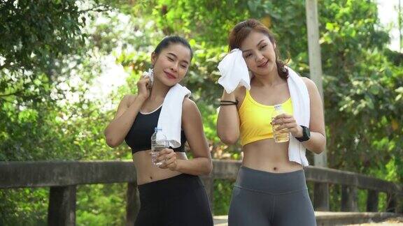 一组两个朋友美丽的亚洲健身女人穿着运动服和毛巾在跑步后喝水休息劳累的女性早上在户外训练后休息擦汗
