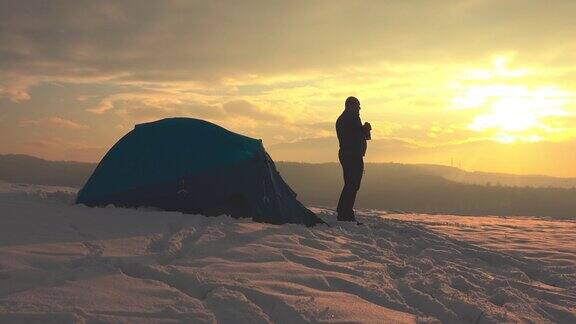 黄昏时分在白雪皑皑的冬季山上穿着短裤的徒步旅行者在帐篷前用保温瓶喝着茶