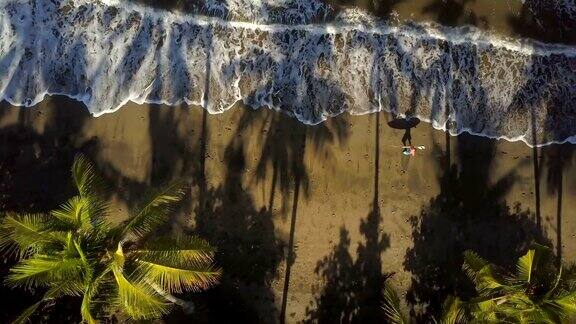 航拍:一个冲浪者背着冲浪板走在沙滩上看着镜头