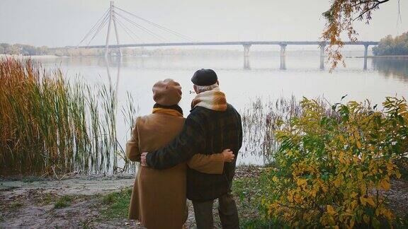 相爱的老夫妇拥抱着微笑着站在河岸上看着跨越它的大桥秋天