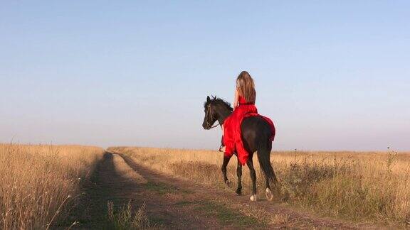 年轻的女孩穿着深红色的长裙骑着黑马穿过干燥的草原