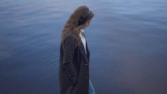 侧面的优雅迷人的年轻女子戴着耳机漫步在河堤上以蓝色的河水为背景自信苗条时尚的白人千禧一代在户外慢动作走路