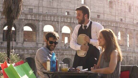 快乐的年轻夫妇游客聊天风趣的意大利优雅的服务员坐在酒吧餐厅前罗马斗牛场在日落开玩笑和大笑有乐趣