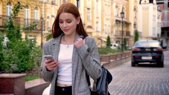 年轻美丽的女人与姜黄色头发走在城市和看着她的手机黄色的建筑背景