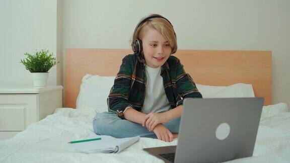 十几岁的男孩使用笔记本电脑和耳机孩子在床上看在线视频课程聊天视频会议男生学习做作业电子学习远程教育课程