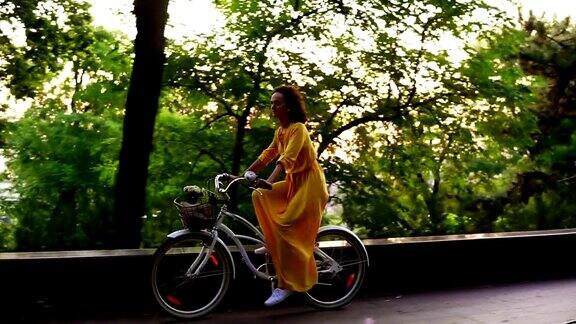 美丽微笑的女人穿着黄色长裙骑着一辆城市自行车里面有一个篮子和鲜花在黎明镜头光晕替身Slowmotion
