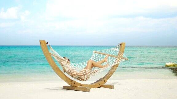 田园诗般的场景美丽的女人日光浴躺在吊床上的海滩蔚蓝的海滩白沙和海蓝宝石的水阳光普照异国风情