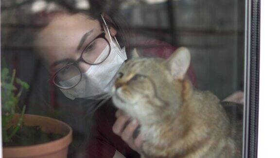 一个戴面具的年轻女子抚摸着一只猫被隔离在家的女孩站在窗口抚摸着那只英国猫玻璃后的景色