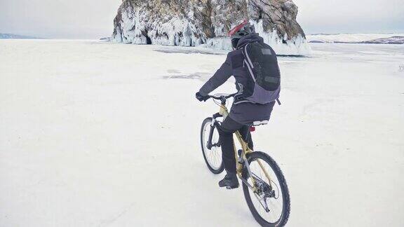 一个人在冰洞附近骑自行车有冰洞和冰柱的岩石非常漂亮骑自行车的人穿着灰色羽绒服背包和头盔旅行者正在骑自行车