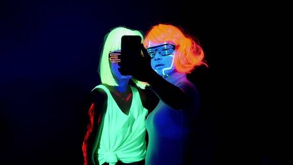 两名性感的网络发光体女性穿着荧光服装在紫外线下使用智能手机自拍