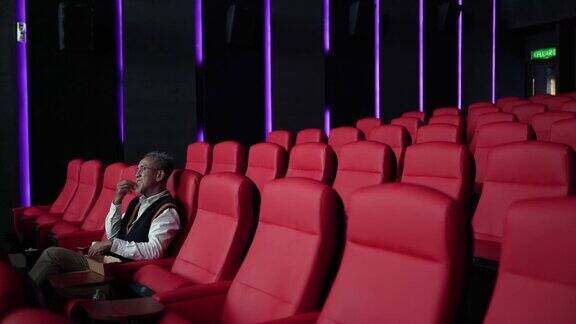一位留着白胡子的亚裔中国男子坐在空荡荡的电影院的红色座位上一边吃一边看电影
