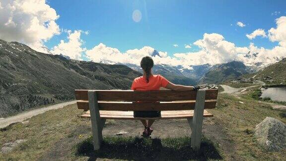 从后面看女徒步旅行者坐在长凳上休息欣赏着山的景色